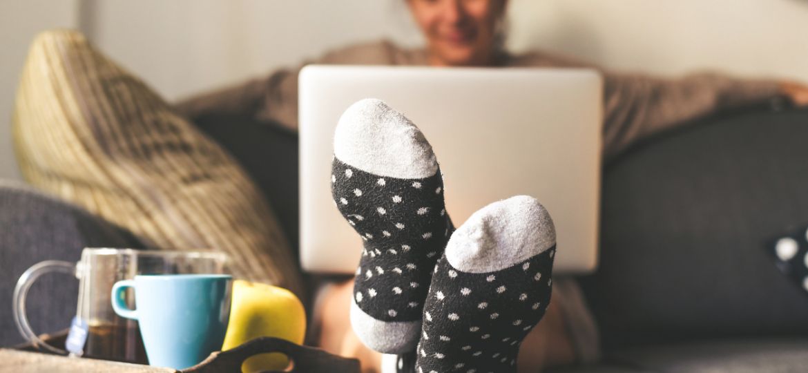 Woman sitting on a couch in the living room with warm socks in a winter morning. Girl using laptop and works at home, having natural breakfast with tea coffee and fruits. Focus on feet in foreground