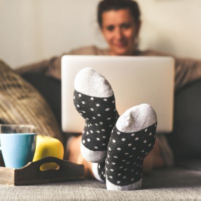 Woman sitting on a couch in the living room with warm socks in a winter morning. Girl using laptop and works at home, having natural breakfast with tea coffee and fruits. Focus on feet in foreground